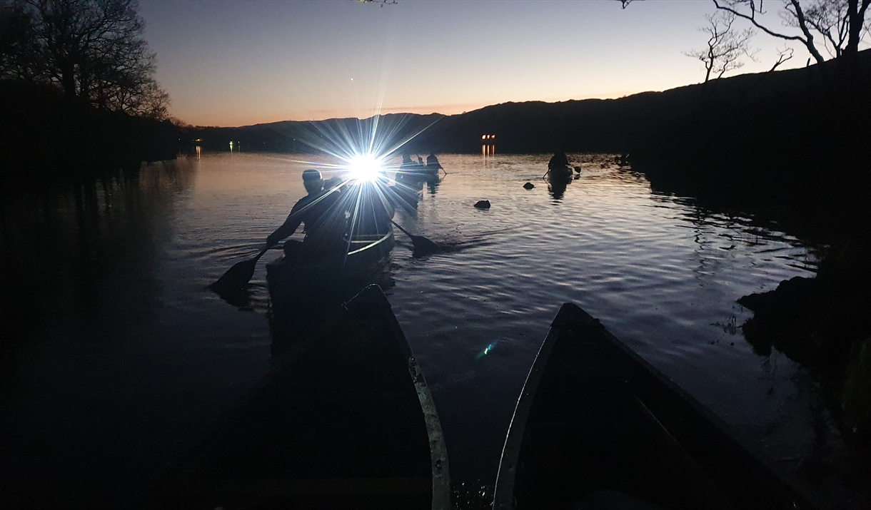 Canoeists on a lake in Cumbria at night, with a bright light from their canoe shining toward the camera, reflecting on the dark water