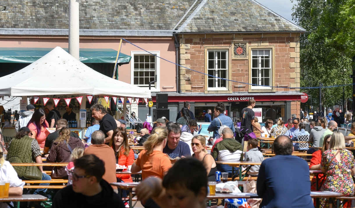 People sat at benches enjoying the International Market in the sunshine with Carlisle's Old Town Hall in the background.