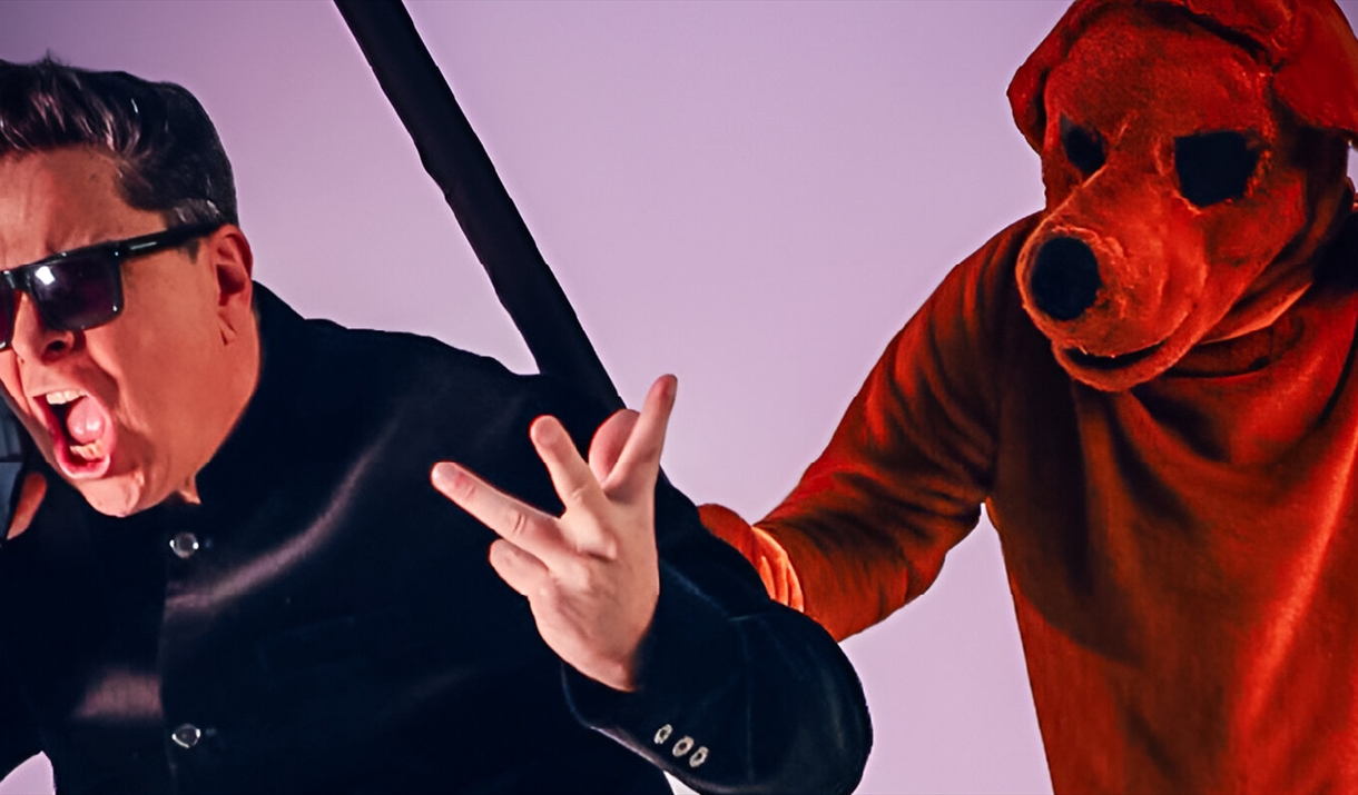 Dom Joly talking on a phone wearing sunglasses, behind him is someone dressed as a red dog