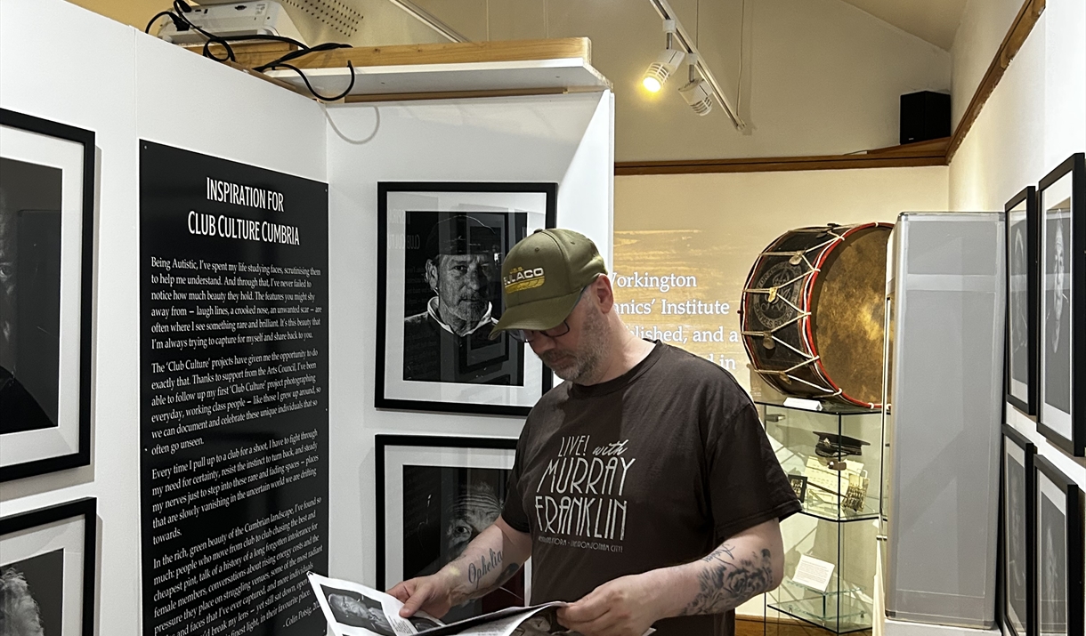 Colin Potsig in The Armitt Museum's exhibition, Club Culture Cumbria. Colin is reading a newspaper and is surrounded by portraits of working men's clu