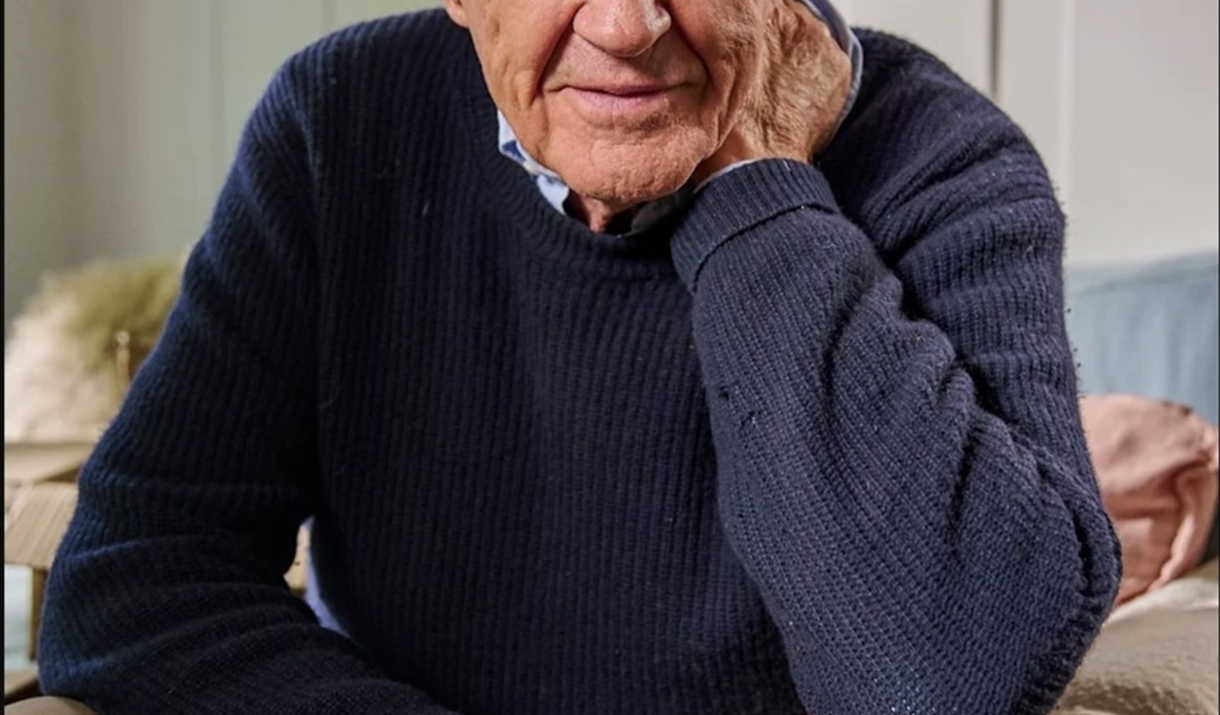 Portrait of a person with white hair wearing a navy blue knitted jumper and beige trousers, seated indoors with one hand resting against the side of t