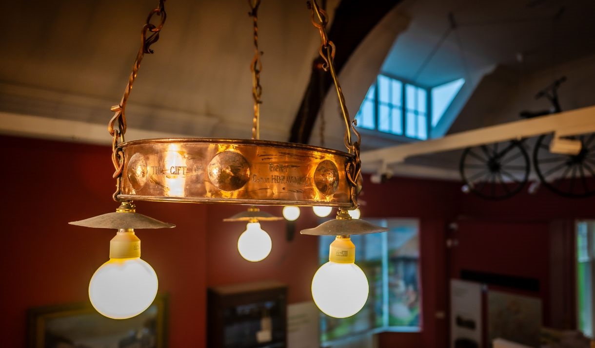 Close-up image of an electrolier (an elaborate late Victorian electric chandelier) on the ceiling of a museum gallery in dim lighting.