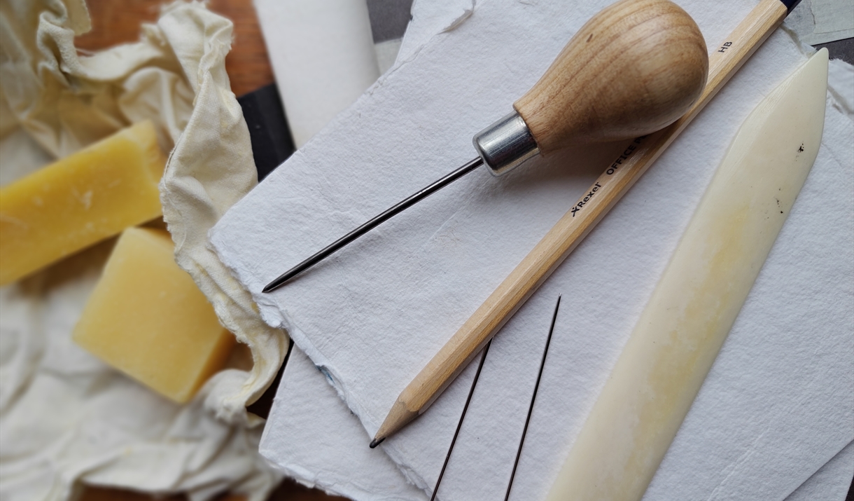 Image shows bookmaking tools such as a pencil, paper, bone folder and a piercing awl
