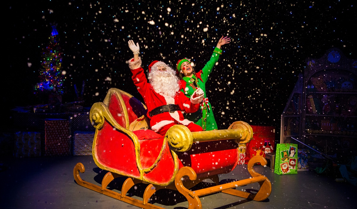 Santa on his sleigh accompanied by an elf both are waving.