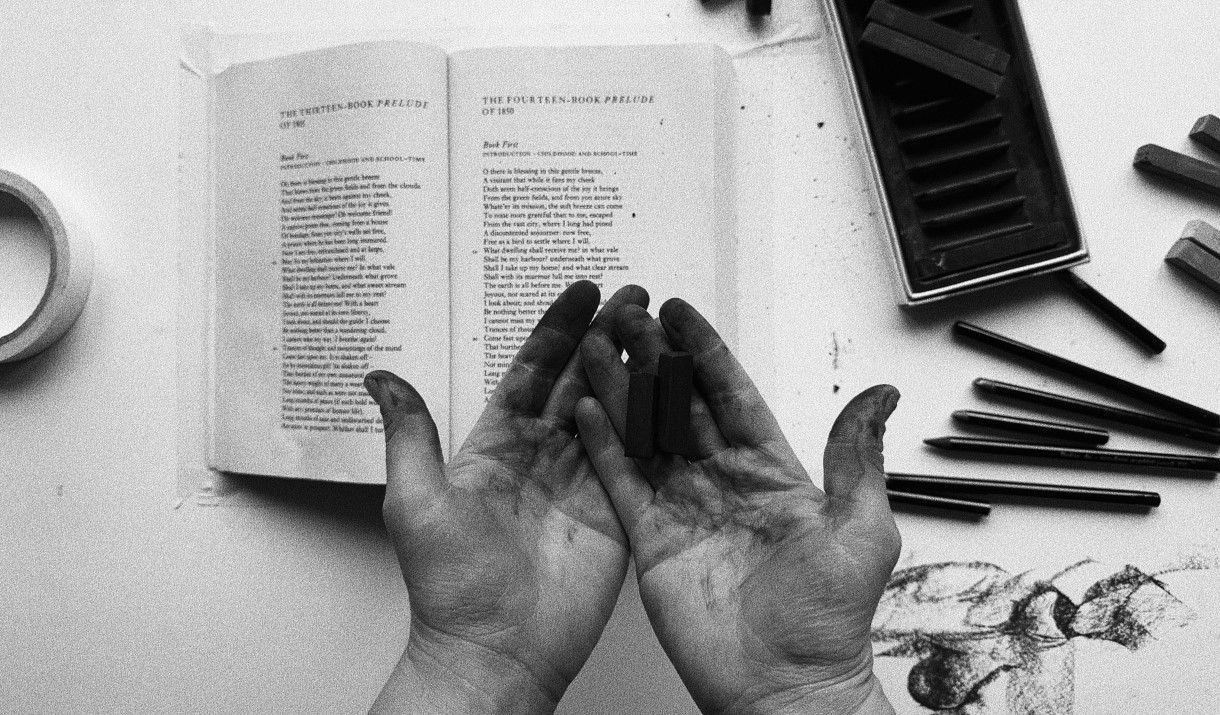 Black and white photo of dirty palms hovering above a table with a book and pencils