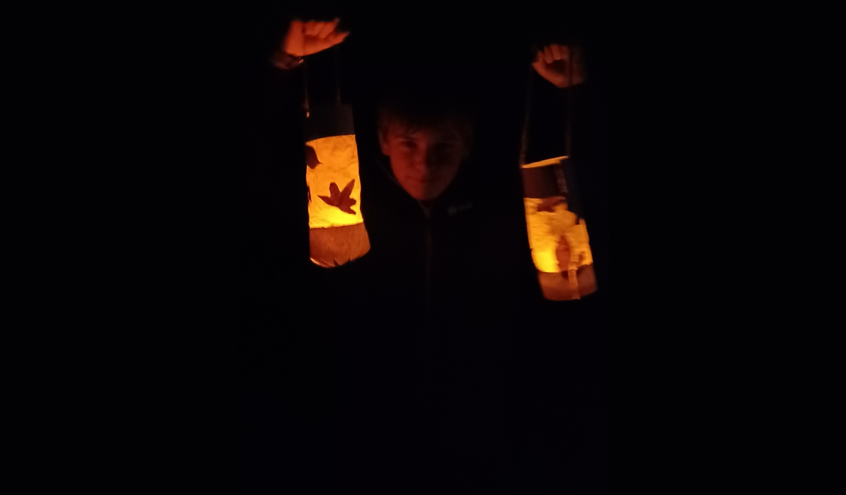A child in a dark setting, illuminated by warm orange light, standing between two glowing lanterns decorated with cut out shapes.