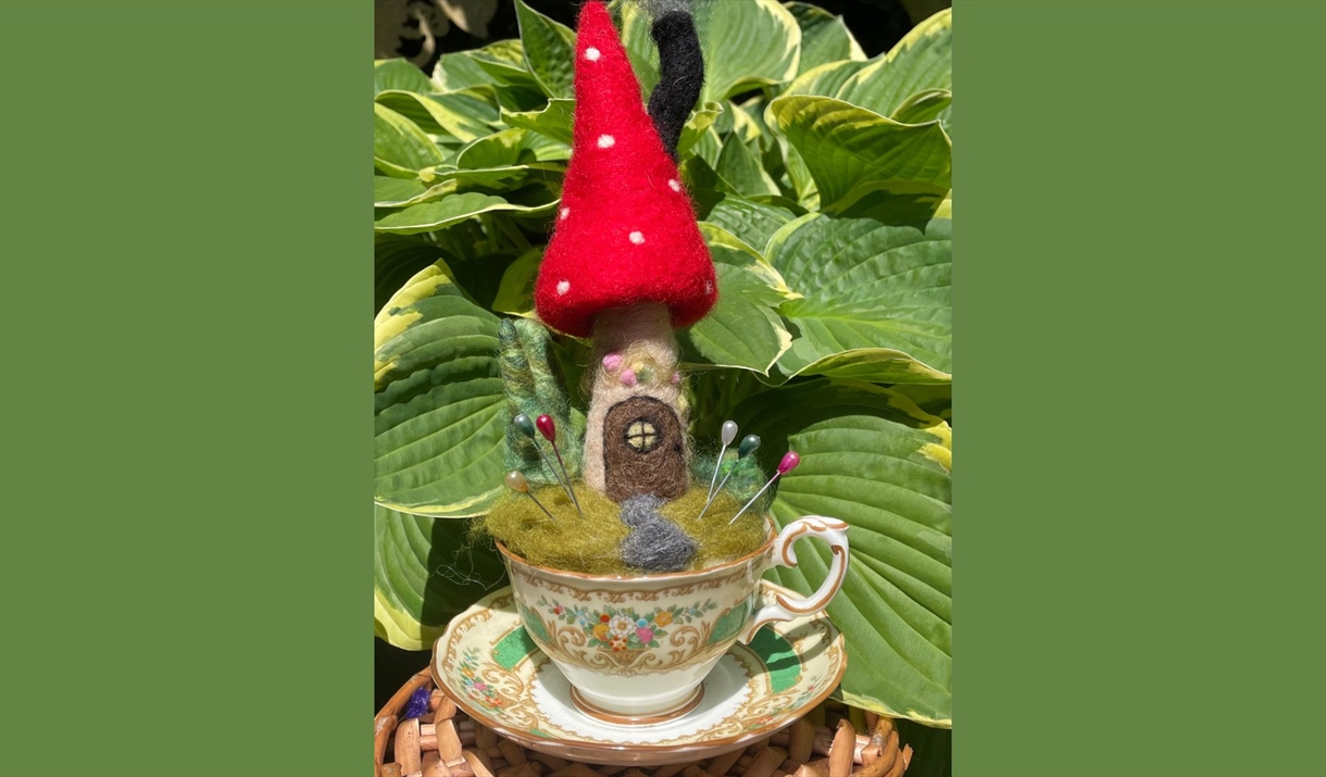 felted mushroom house pincushion with a red cap and white spots, set in a vintage teacup with colorful pins, placed outdoors against a backdrop of lar
