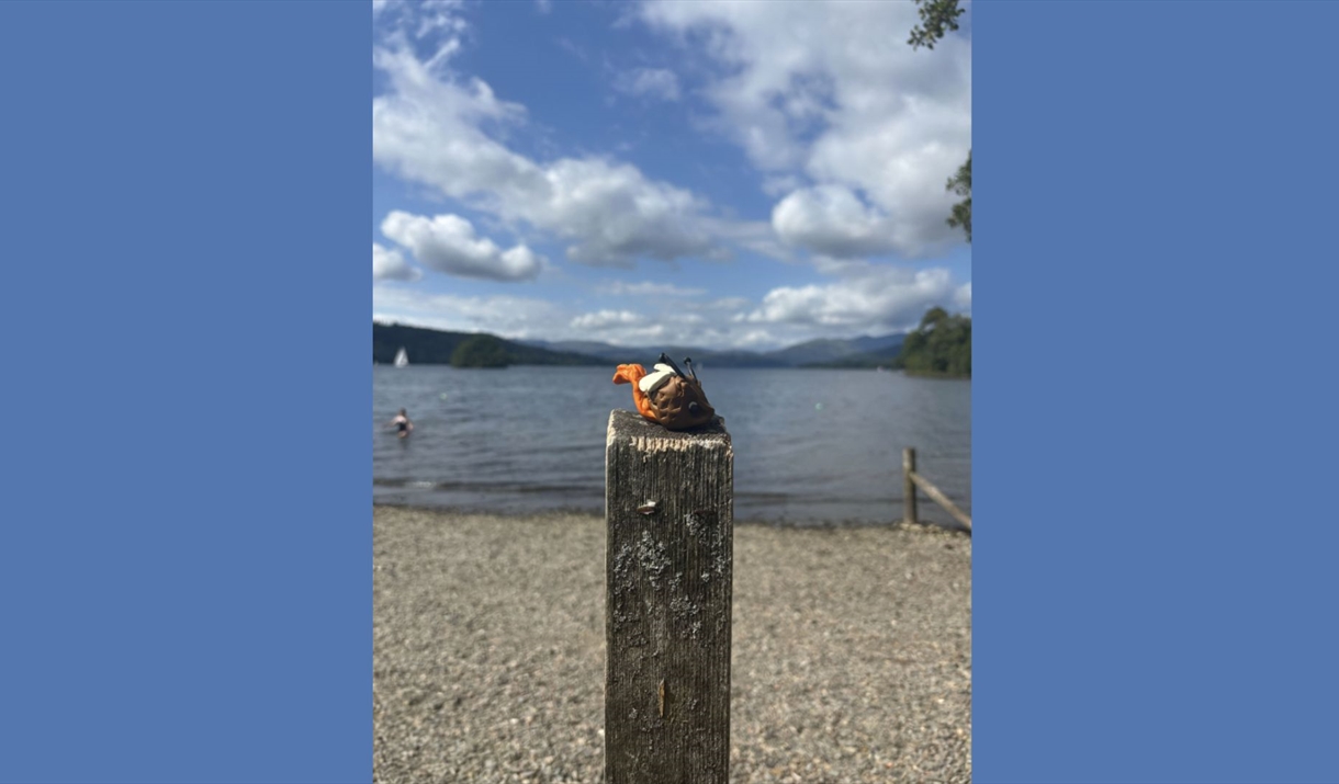 Monster made of clay seated on a wooden pole with a backdrop of lake Windermere.