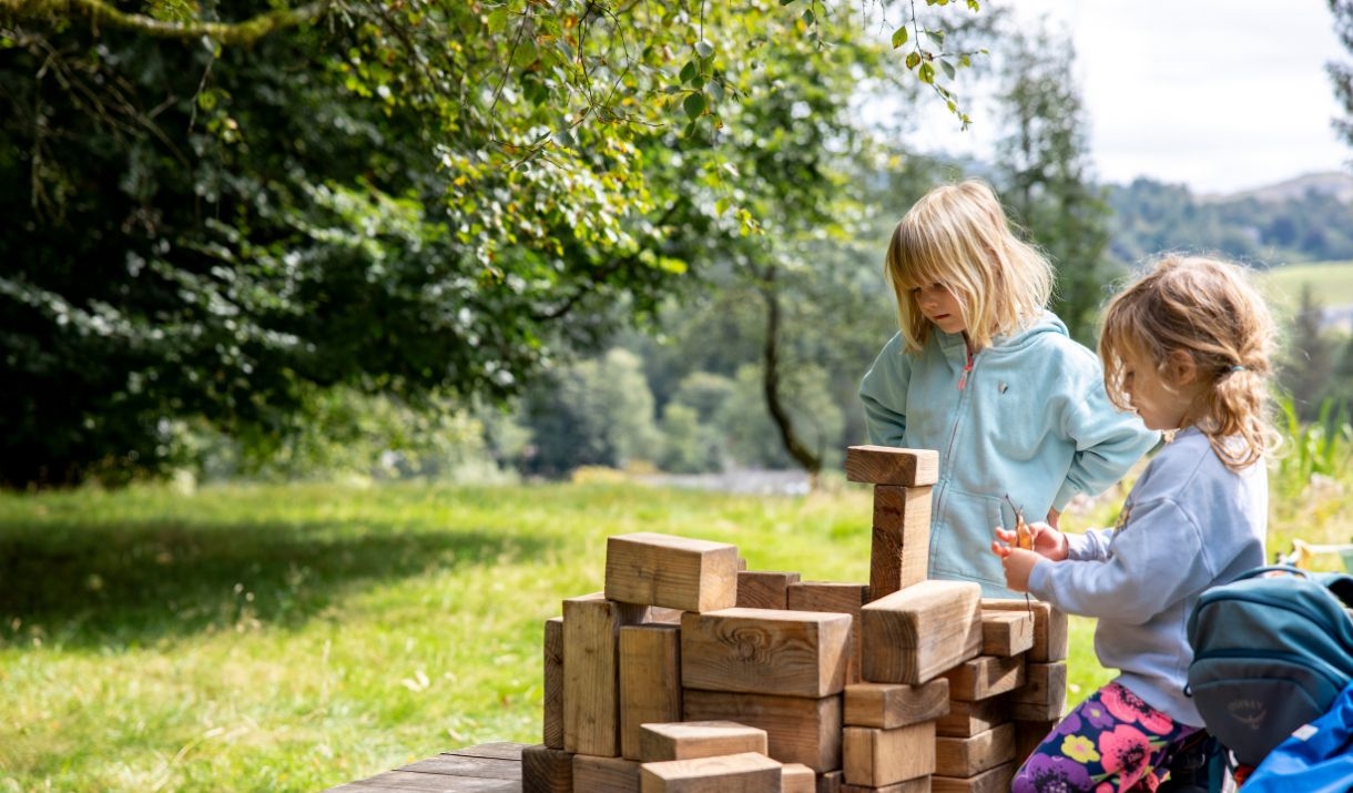 Two children playing with large wooden blocks outside on a sunny day