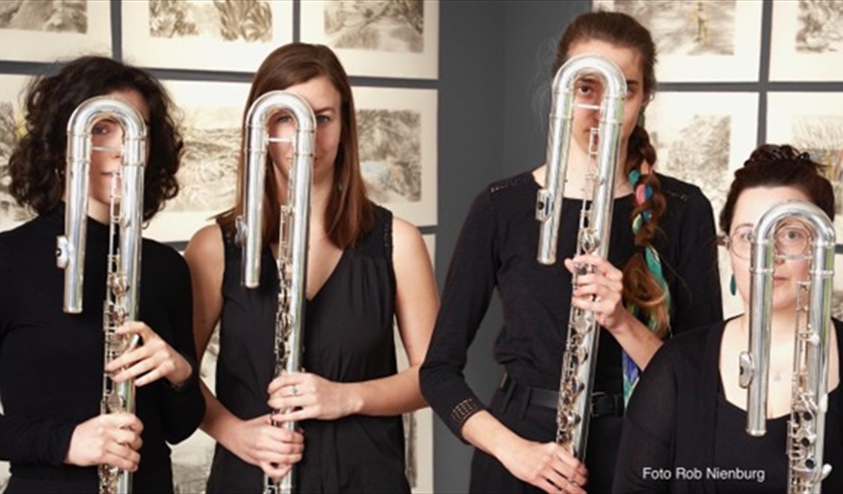 Four women dressed in black stand holding large U-shaped flutes (contrabass flutes) in front of their faces, partially obscuring them. They are standi