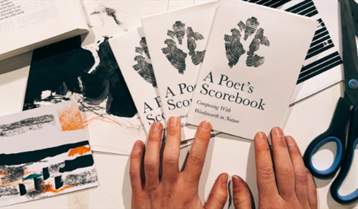 Hands arranging three booklets titled A Poet’s Scorebook on a desk with scissors, a ruler, an open book, and abstract artwork scattered around.