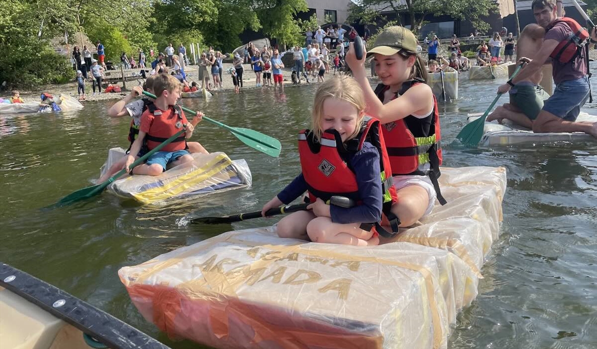 Children in life jackets paddle small rafts on a lake as people watch from the shore.