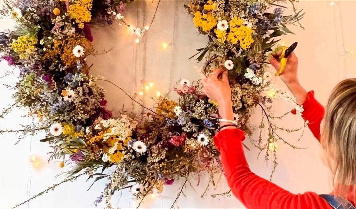 A person wearing a red long-sleeve shirt adjusts a large floral wreath made of colorful dried flowers, including yellow, white, and purple blooms, han