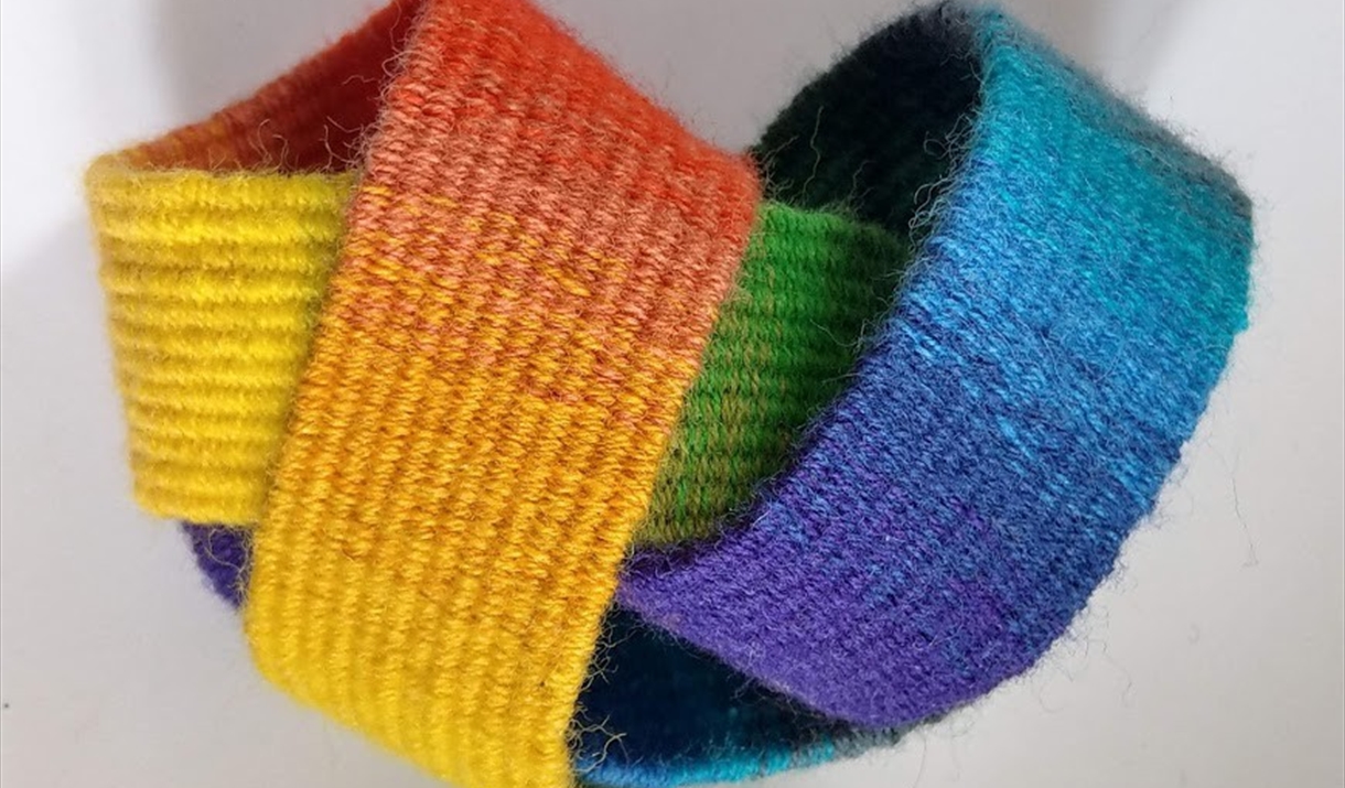 A rainbow-colored weaving displayed on a white surface, featuring vibrant hues and a soft, flowing texture.