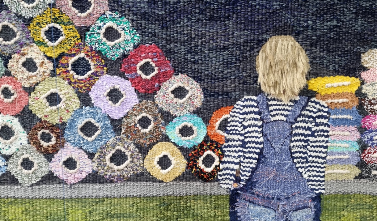 Tapestry piece of a women in dungarees looking at lots of different colour Yarn