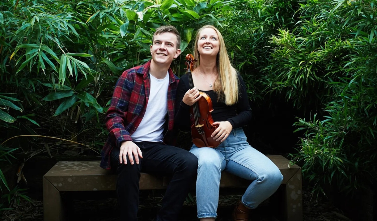 a young man and woman sitting on a wooden bench surrounded by dense green foliage