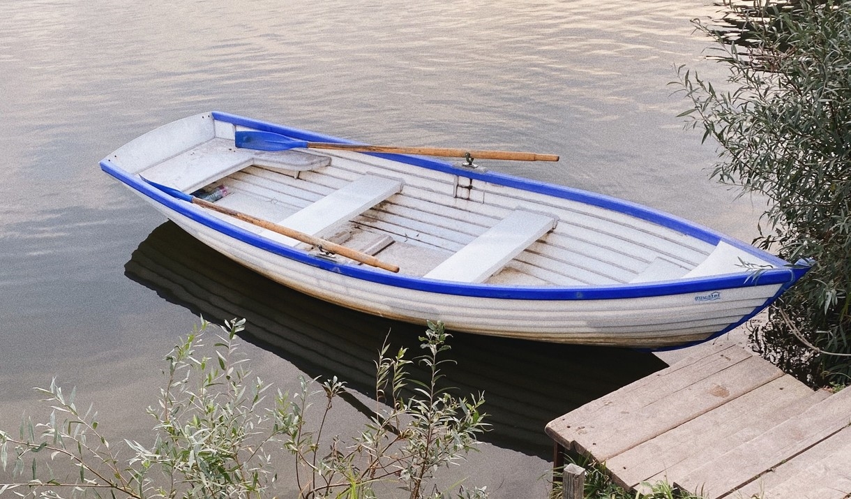 A white rowing boat with blue trim and wooden oars moored by a small jetty on a calm lake