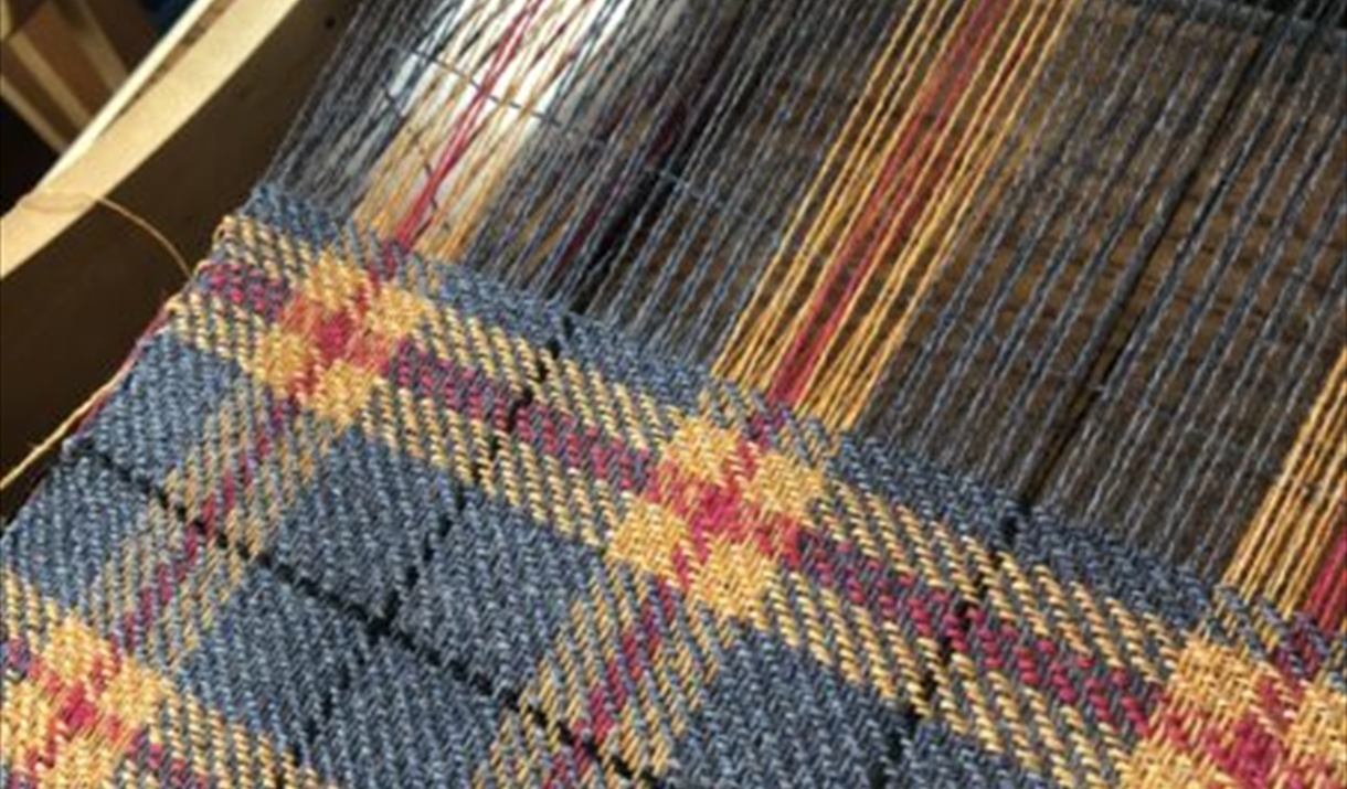 Close-up of a woven tartan fabric on a loom, featuring a pattern of blue, yellow, and red threads in a traditional plaid design