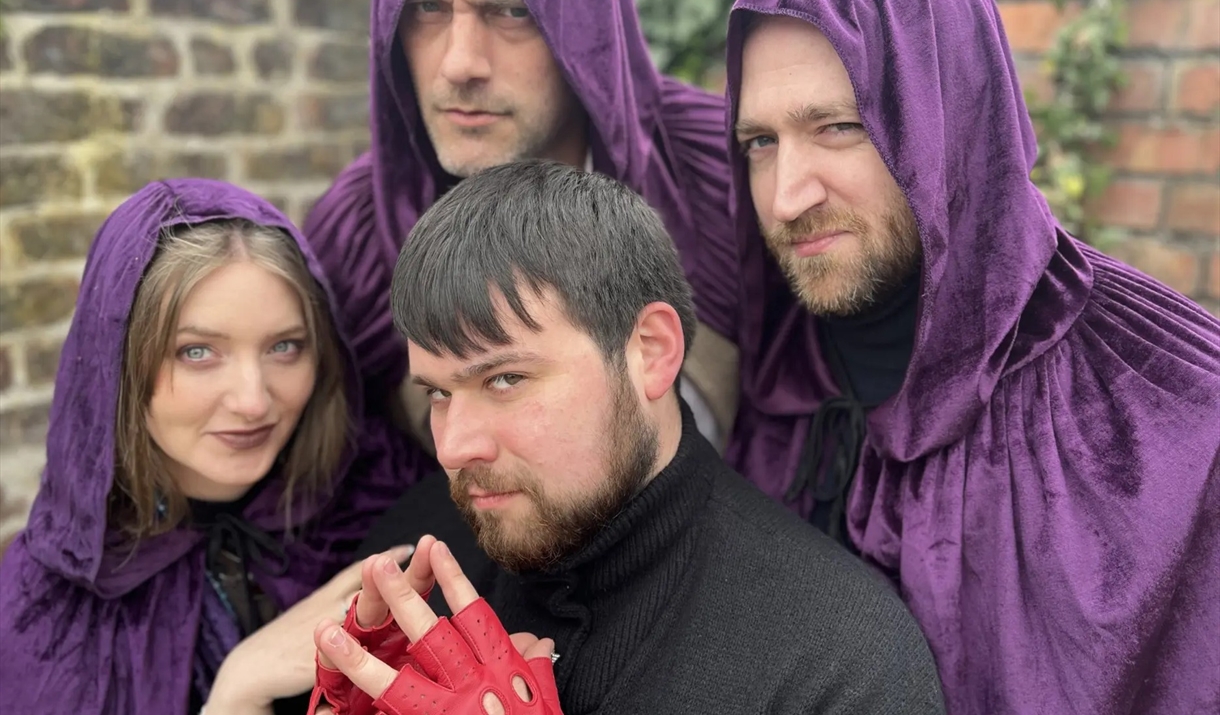 There are four people posing together. Three of them are wearing deep purple hooded cloaks the fourth is in black wearing red gloves