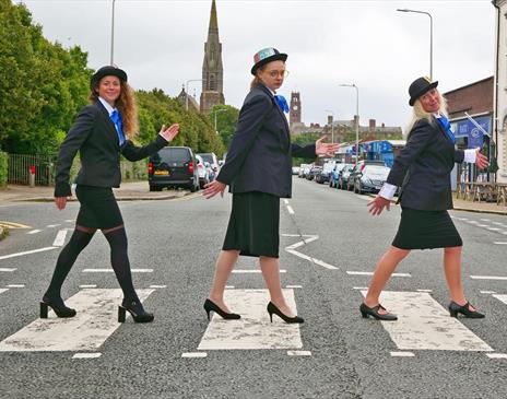 Three women dressed in black suits, blue ties, and bowler hats walking across a zebra crossing in a humorous recreation of the Beatles' Abbey Road pho