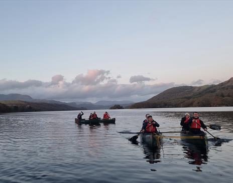 8 canoeists paddling on a calm lake at dusk in Cumbria, silhouetted against a soft, fading sky with hills in the distance