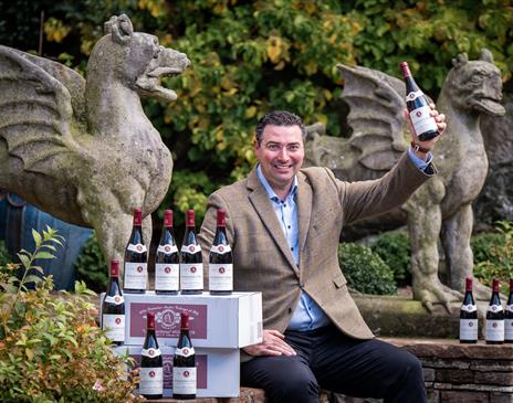 A smiling man holding up a bottle of red wine sat next to two stone statues