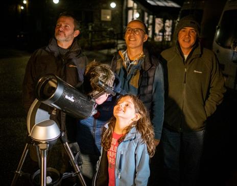 Two children looking through a telescope at night accompanied by 4 adults