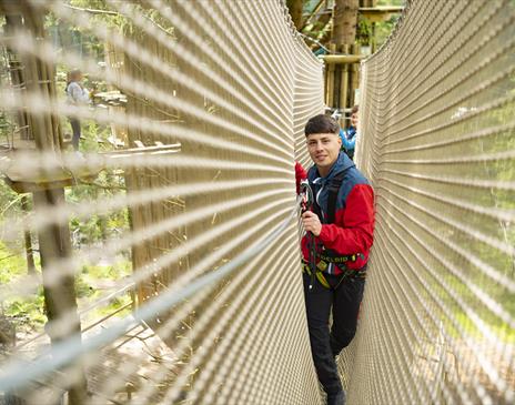 A young man wearing a red and blue jacket and safety harness navigating a rope bridge in a treetop adventure course, surrounded by lush green forest.