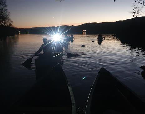 Canoeists on a lake in Cumbria at night, with a bright light from their canoe shining toward the camera, reflecting on the dark water