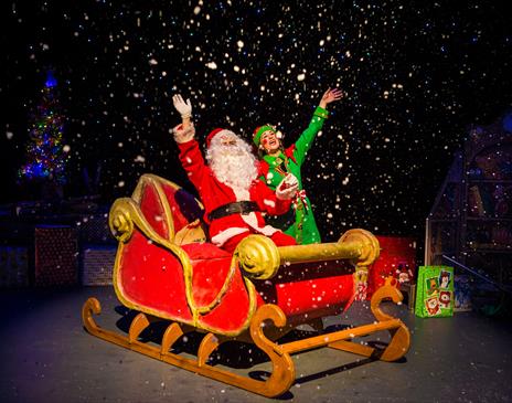 Santa on his sleigh accompanied by an elf both are waving.