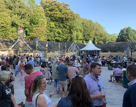 Large crowd gathered at an outdoor event with livemusic, food, and drinks. Set against a backdrop of historic stone buildings and trees on a sunny day
