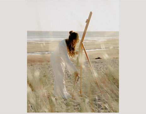Women on the beach setting up an easel dressed in all white.