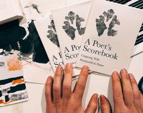 Hands arranging three booklets titled A Poet’s Scorebook on a desk with scissors, a ruler, an open book, and abstract artwork scattered around.