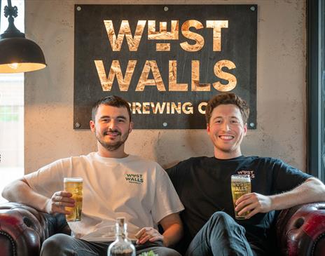 Founders of West Walls Brewing Co, seated inside the brewery, holding beers in Carlisle, Cumbria