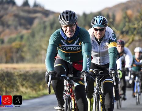 Group of cyclists riding on a country road, with two riders in green jerseys leading the way and smiling.