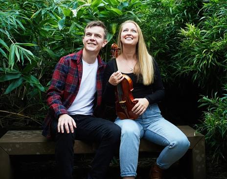 a young man and woman sitting on a wooden bench surrounded by dense green foliage