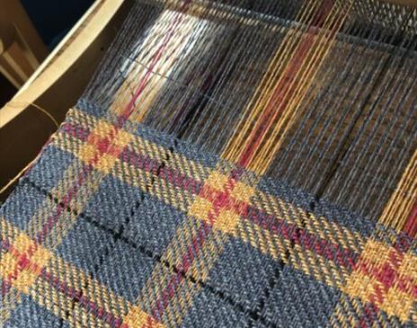 Close-up of a woven tartan fabric on a loom, featuring a pattern of blue, yellow, and red threads in a traditional plaid design