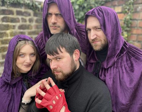 There are four people posing together. Three of them are wearing deep purple hooded cloaks the fourth is in black wearing red gloves