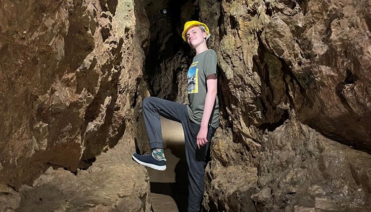 A teenage boy standing in a narrow underground tunnel, leaning against the wall and resting his foot on the rock in front of him.