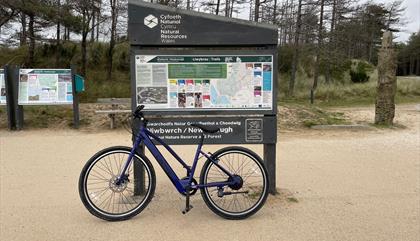 Photos of bike leaning against signage in Newborough Forest of the walking and cycle routes,