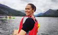 A woman, wearing a red buoyancy aid, turns around to smile at the camera, her joy evident against the breathtaking backdrop of Mount Snowdon at Llyn P