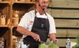 Graham Tinsley, MBE, doing a cooking demonstration in the demonstration kitchen with jars on a shelf behind him and he's whisking ingredients in a met
