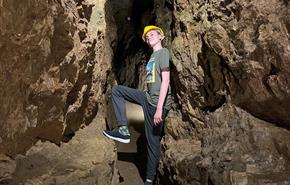 A teenage boy standing in a narrow underground tunnel, leaning against the wall and resting his foot on the rock in front of him.