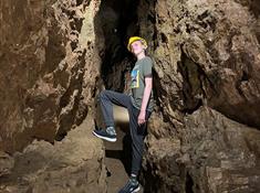 A teenage boy standing in a narrow underground tunnel, leaning against the wall and resting his foot on the rock in front of him.