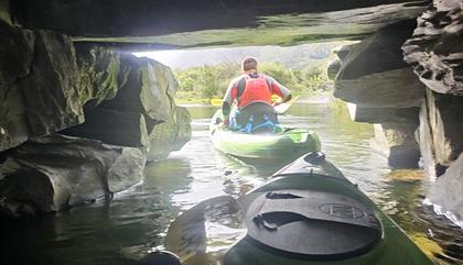 Adventurous group navigating through a shadowy tunnel in green sit-on-top kayaks on Llyn Padarn Lake, North Wales. The kayakers, wearing red buoyancy