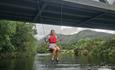 A woman in a red buoyancy aid swings joyfully on a rope below a train bridge at Llyn Padarn Lake, North Wales. The background features lush greenery a