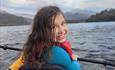 A young girl in a red buoyancy aid turns to smile brightly at the camera, her happiness radiating against the majestic Mount Snowdon at Llyn Padarn La