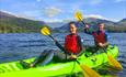 A couple paddles together in a green sit-on-top kayak on Llyn Padarn Lake, with the majestic Mount Snowdon towering in the background under a clear bl