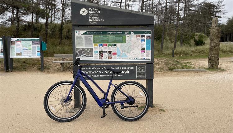Photos of bike leaning against signage in Newborough Forest of the walking and cycle routes,