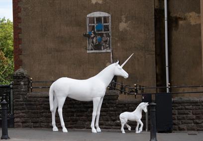 2 large white unicorn statues stood in front of Banksy graffeti
